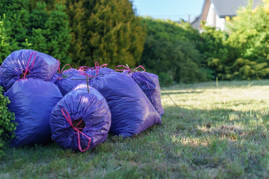 A group of seven large purple plastic rubbish bags, tightly sealed with pink ties, are stacked closely together on a grassy area, partially leaning against a green bush on the left side. The bags appear to contain waste, with some showing slight bulges and irregular shapes, indicating they are filled. The grass beneath them is uneven, with patches of dry and fresh grass visible, and the bags cast soft shadows on the ground. In the background, there is a lush, dense hedge and a row of trees with green foliage, along with the rooftops and upper structures of nearby residential houses, suggesting a suburban setting. The scene is illuminated by natural sunlight, creating a bright and clear atmosphere characteristic of a sunny day. The overall environment suggests an area prepared for waste removal or collection, aligning with private or independent rubbish disposal services, such as those offered by Rubbish Clearance Finchley.