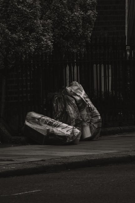 Two large black plastic rubbish bags filled with waste are positioned on the edge of a paved sidewalk, with some appearing more crumpled and overstretched, indicating they are full. The bags have a glossy texture, reflecting some minimal ambient light, and display printed markings, though the text is partly obscured and not fully readable. They are placed directly against a black metal fence with vertical bars, situated in a residential or urban environment. Behind the fence, there are dense shrubbery and part of a brick building or structure, with the foliage extending upward and providing a contrasting background to the bags. The scene is lit in subdued, natural light, suggesting either an overcast day or shaded area, emphasizing the dark, muted tones throughout the image. The arrangement and condition of the rubbish bags imply they are awaiting collection or disposal, typical of independent waste handling or private rubbish clearance services, such as those provided by Rubbish Clearance Finchley, operating away from formal council collection systems.