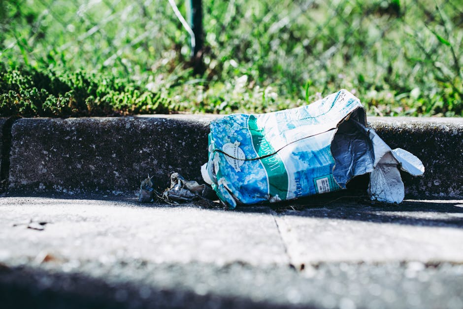 A group of seven large purple plastic rubbish bags, tightly sealed with pink ties, are stacked closely together on a grassy area, partially leaning against a green bush on the left side. The bags appear to contain waste, with some showing slight bulges and irregular shapes, indicating they are filled. The grass beneath them is uneven, with patches of dry and fresh grass visible, and the bags cast soft shadows on the ground. In the background, there is a lush, dense hedge and a row of trees with green foliage, along with the rooftops and upper structures of nearby residential houses, suggesting a suburban setting. The scene is illuminated by natural sunlight, creating a bright and clear atmosphere characteristic of a sunny day. The overall environment suggests an area prepared for waste removal or collection, aligning with private or independent rubbish disposal services, such as those offered by Rubbish Clearance Finchley.