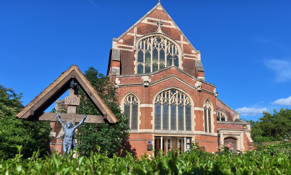 A historic red-brick church with large pointed arched stained glass windows and ornate stone detailing, set against a vivid blue sky. In the foreground, a wooden crucifix with a metal figure of Jesus Christ is partially covered by green foliage, including bushes and small trees. The church's stone and brick exterior showcases a mix of smooth and rough textures, with the brickwork arranged in a decorative pattern, and the windows featuring intricate tracery. The scene appears to be taken from a garden or landscaped area surrounding the church, with bright sunlight casting shadows and illuminating the building’s architectural features. This setting emphasizes the church’s prominent position within a peaceful environment, suitable for quiet reflection or private service, aligning naturally with themes of churchyard maintenance and the potential for on-site rubbish clearance or preservation of the surrounding greenery, as handled by services like Rubbish Clearance Finchley.