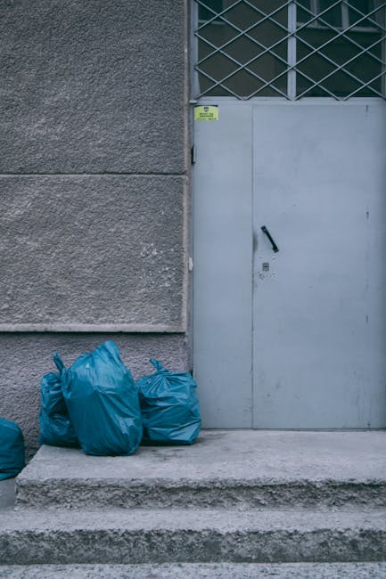 The image depicts four blue rubbish bags placed on a concrete step outside a building, positioned to the left of a closed grey metal door with a black handle and a small lock. The building's exterior wall is textured with a rough, dark grey surface, and to the right of the door, there is a diagonal lattice security grille covering a small window or ventilator, also painted grey. The ground in front of the door appears to be a concrete pavement, with a slight step leading up to the entrance. The scene suggests a typical urban setting where waste is temporarily stored outside, possibly for collection by a private rubbish removal service such as Rubbish Clearance Finchley, highlighting an example of on-site waste disposal suitable for businesses or residential properties seeking alternative rubbish management solutions. The lighting is natural, illustrating an overcast day, with no other objects or people visible in the scene.
