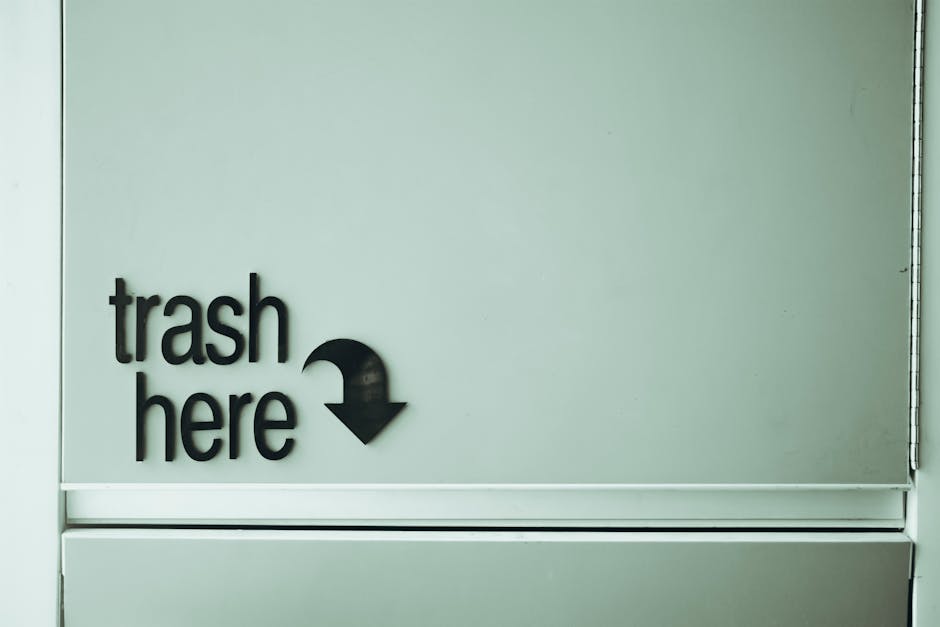 A close-up image of a minimalist wall-mounted sign with the words 'trash here' in black lowercase letters on a light grey or pale green background. Next to the text is a downward-pointing black arrow, indicating the location of a trash receptacle or bin, which is not visible in the image. The sign appears to be affixed to a smooth, flat surface, possibly part of a waste disposal station or designated rubbish collection point in an indoor setting. The environment looks clean and well-maintained, suggesting an organized area for waste disposal, aligning with professional rubbish removal or private waste handling services such as those offered by Rubbish Clearance Finchley. The lighting is even, with no shadows or reflections, highlighting the clarity and simplicity of the sign used to direct waste disposal activities in a systematic manner.