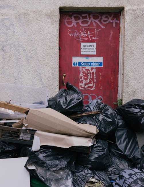The image depicts four blue rubbish bags placed on a concrete step outside a building, positioned to the left of a closed grey metal door with a black handle and a small lock. The building's exterior wall is textured with a rough, dark grey surface, and to the right of the door, there is a diagonal lattice security grille covering a small window or ventilator, also painted grey. The ground in front of the door appears to be a concrete pavement, with a slight step leading up to the entrance. The scene suggests a typical urban setting where waste is temporarily stored outside, possibly for collection by a private rubbish removal service such as Rubbish Clearance Finchley, highlighting an example of on-site waste disposal suitable for businesses or residential properties seeking alternative rubbish management solutions. The lighting is natural, illustrating an overcast day, with no other objects or people visible in the scene.