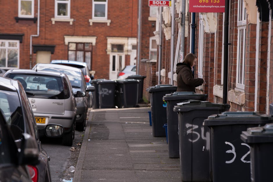 A residential street scene featuring a row of black wheelie bins positioned along the pavement outside brick-built terraced houses. The bins are made of plastic with lids that are mostly closed, some showing signs of wear with graffiti markings in white paint on their sides. The pavement is concrete, with a narrow curb separating it from the road where several parked cars are visible, including a small silver hatchback and a few other vehicles further down the street. The background displays brick houses with white-framed windows and decorative stone lintels. A woman dressed in a dark coat stands near one of the bins, possibly preparing to dispose of rubbish. Overhead, a red sign hangs from one of the houses, indicating a private property or notice. The scene is lit with natural daylight, capturing an everyday urban environment where independent waste disposal aligns with local rubbish collection practices, and the presence of numerous bins suggests a focus on domestic waste management efforts.