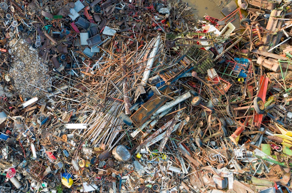 A large, densely packed pile of mixed metal, plastic, and wooden debris of various shapes and sizes, including rusted metal pipes, wire frames, plastic containers, and broken machinery components, with some items showing corrosion and weathering. The debris is scattered across a gravel surface, with some pieces leaning or overlapping others. In the background, a muddy area with water and some green vegetation is visible. The overall scene depicts a scrapyard or dump site for waste sorting and recycling, indicative of private waste handling services such as rubbish clearance provided by companies like Rubbish Clearance Finchley, which specialize in managing bulk waste and scrap removal. The lighting is natural, with diffuse sunlight illuminating the chaotic jumble of materials, highlighting the textures of rusted metals, plastic, and organic matter.
