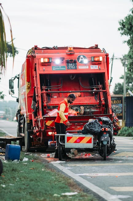 A worker wearing a yellow and red uniform is loading waste into the rear chamber of a large red rubbish collection truck, which is parked on the side of a road. The truck's rear door is open, revealing a metal interior used for compacting waste. The worker is standing next to a small metal platform with a black plastic bag positioned nearby on top of the platform. The area around the truck includes a patch of grass and a concrete curb, with some scattered debris on the ground. In the background, there are trees, utility poles with overhead wires, and a signboard partially visible on a building. Overcast sky and soft natural lighting suggest daytime conditions. This scene illustrates independent waste collection likely related to private rubbish removal services, emphasizing on-site clearance of household waste as performed by a waste management professional.
