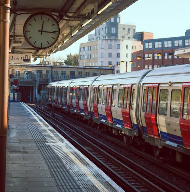 The image depicts a London Underground train with a silver body featuring red and blue stripes parked at a station platform. The train has multiple carriages with large windows, and the exterior appears clean and well-maintained. The station platform has tactile paving strips near the edge for passenger safety, and the surface is made of concrete or similar material with visible seams and minor texture. Overhead, an overhanging structure supports lighting fixtures and a large round clock with a white face, black hour and minute hands, and red second hand, showing the time as approximately 1:55. In the background, there are various modern multi-storey buildings with a mix of white, grey, and brown facades, and a partly cloudy sky is visible. The scene is well-lit, suggesting daytime, and was likely captured during a period of regular train services, aligning with the context of routine public transport and potentially independent or non-publicly-operated service aspects relevant to waste and rubbish removal environments like those managed by Rubbish Clearance Finchley.