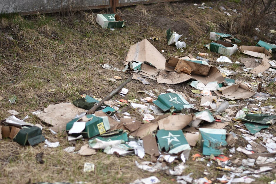 A patch of ground with scattered debris and litter, including numerous flattened cardboard boxes in various shades of brown and green, some with printed logos and labels, lying on grass and dirt. The cardboard appears torn and crumpled, with some partially unfolded or overlapping each other. Small pieces of paper, plastic wrappers, and other miscellaneous trash are dispersed around the larger cardboard pieces. In the background, there is a wooden fence or barrier, partially visible, with a weathered surface. The environment is outdoors, and the scene is illuminated by natural daylight, creating a neutral, unobtrusive atmosphere. This image illustrates common instances of debris left on site, relevant to private waste clearance, such as rubbish removal services, including those provided by Rubbish Clearance Finchley, and highlights the need for efficient rubbish collection to maintain cleanliness.