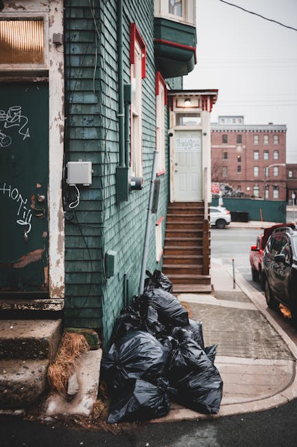 A close-up view of the exterior of a residential building painted in greenish-blue wooden siding, with a small set of wooden stairs leading to a white door at the top. Several black rubbish bags are piled on the sidewalk next to the building, partially resting on a small patch of snow and mud at the base of the wall. The scene is set on a paved street with parked cars, including a dark-colored vehicle directly beside the rubbish bags. In the background, there is an urban landscape with brick buildings, utility wires overhead, and an overcast sky. The presence of rubbish bags suggests recent waste removal activity, possibly part of private rubbish clearance services such as those offered by Rubbish Clearance Finchley, highlighting an outdoor setting where alternative waste handling methods are employed. The condition of the building’s exterior, along with the rubbish bags' placement, illustrates typical scenes of residential waste disposal in an urban environment focused on waste collection and clearance.
