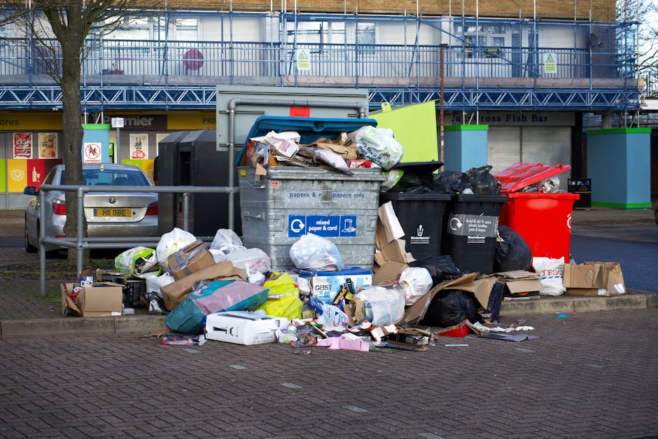 A patch of ground with scattered debris and litter, including numerous flattened cardboard boxes in various shades of brown and green, some with printed logos and labels, lying on grass and dirt. The cardboard appears torn and crumpled, with some partially unfolded or overlapping each other. Small pieces of paper, plastic wrappers, and other miscellaneous trash are dispersed around the larger cardboard pieces. In the background, there is a wooden fence or barrier, partially visible, with a weathered surface. The environment is outdoors, and the scene is illuminated by natural daylight, creating a neutral, unobtrusive atmosphere. This image illustrates common instances of debris left on site, relevant to private waste clearance, such as rubbish removal services, including those provided by Rubbish Clearance Finchley, and highlights the need for efficient rubbish collection to maintain cleanliness.